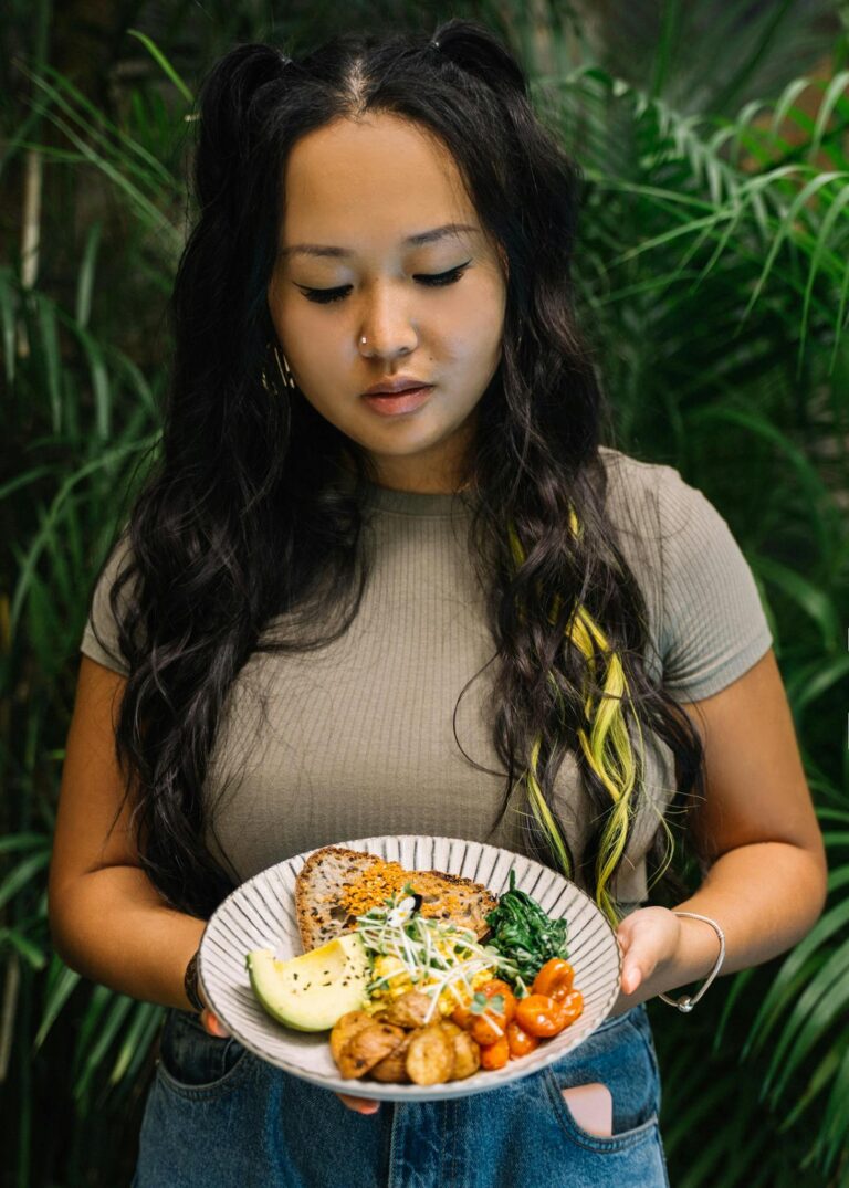 Asian woman holding a plate of healthy food with vegetables and bread, surrounded by lush greenery indoors.