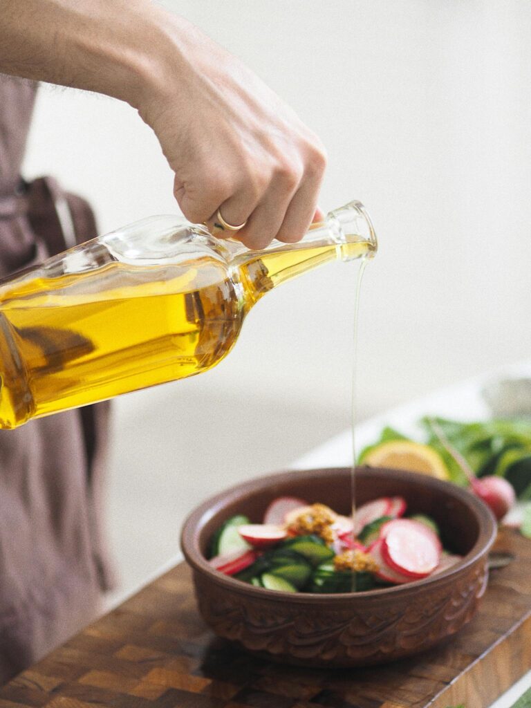 Hand pouring olive oil over a fresh vegetable salad in a kitchen setting.