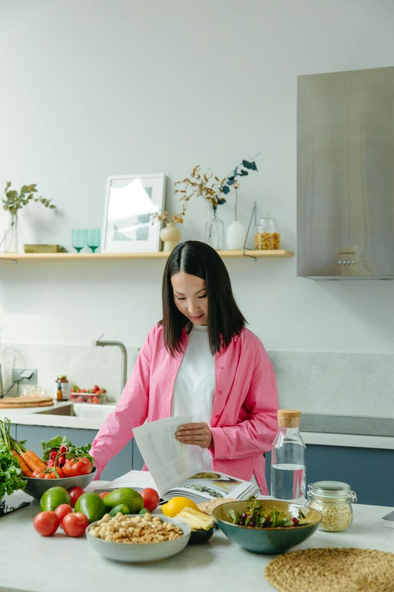 Woman in pink shirt preparing fresh vegetables and reading a recipe in a modern kitchen.